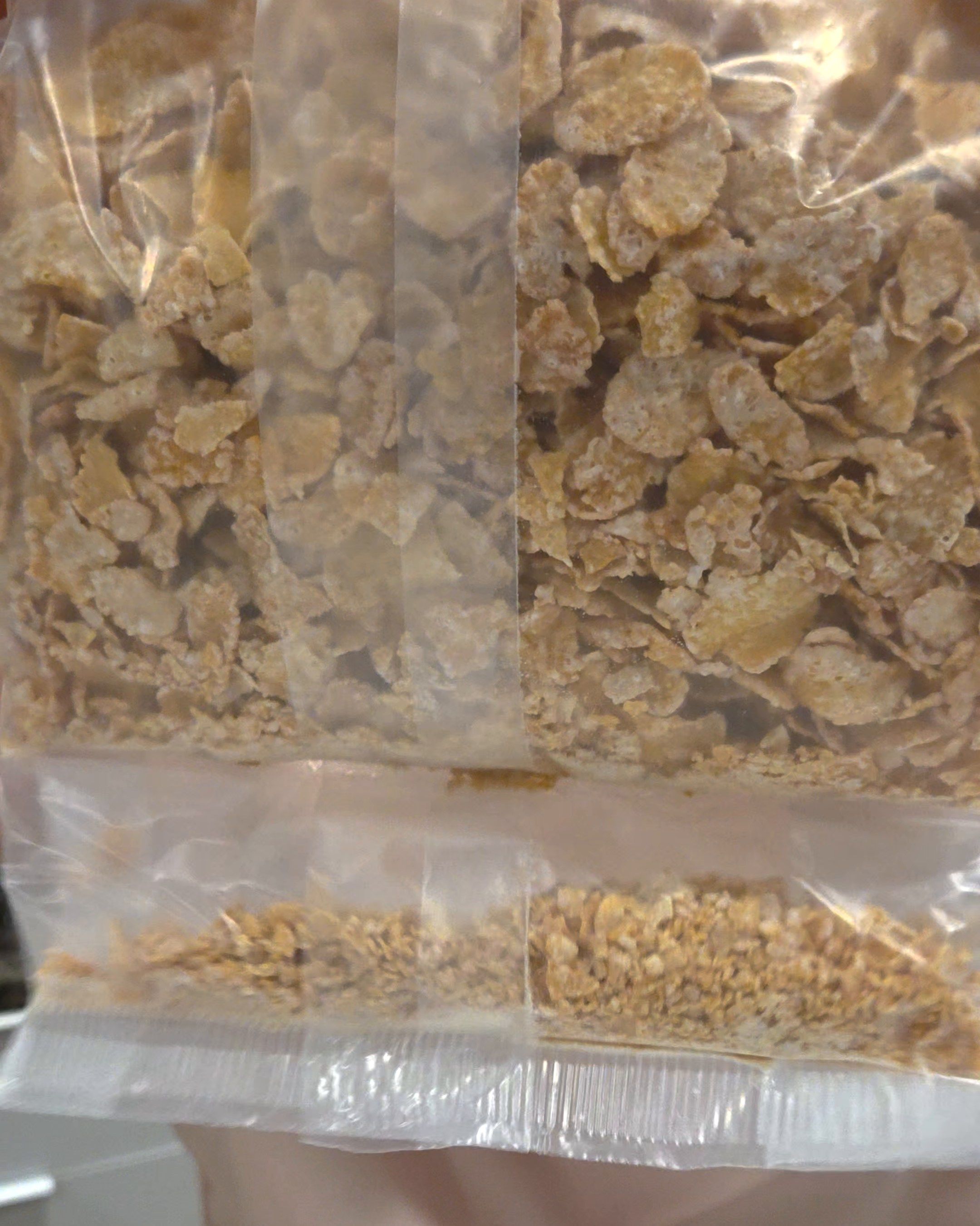 Close-up of a clear bag of cereal flakes. Below a seal near the bottom, a narrow compartment holds a layer of small crumbs.